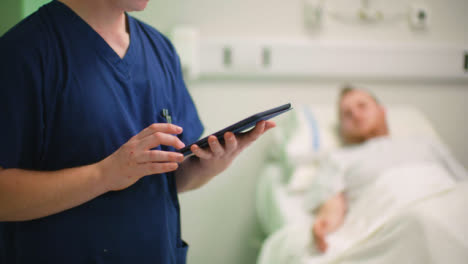 Male-Nurse-by-Patient-Making-Notes-on-Tablet
