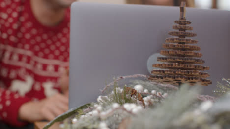 Extreme-Close-Up-of-Christmas-Table-Decorations-In-Front-of-Couple-Using-a-Laptop-