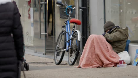 Long-Shot-of-Homeless-Person-Sitting-On-Ground-On-Busy-Street-In-Oxford-England
