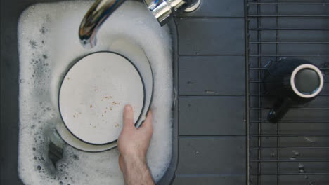 Top-Down-Shot-of-Male-Hands-Placing-Dirty-Dishes-into-Sink-of-Soapy-Water