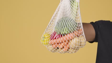 Mid-Shot-of-Man-Holding-up-Bag-of-Vegetables-In-Front-of-Yellow-Background