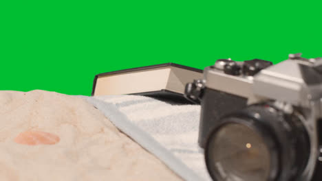 Summer-Holiday-Concept-Of-Book-Camera-Beach-Towel-On-Sand-Against-Green-Screen