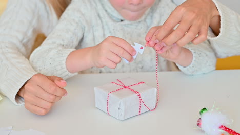 Child-Decorates-The-Bow-Of-A-Wrapped-Christmas-Gift-With-A-Paper-Tree