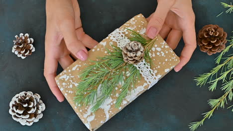 Female-Hands-Shows-A-Wrapped-Gift-Decorated-With-A-Pine-Cone,-A-Branch-And-Painted-Snow-On-A-Table