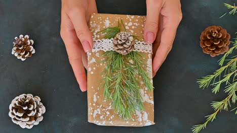 Female-Hands-Shows-A-Wrapped-Gift-Decorated-With-A-Pine-Cone,-A-Branch-And-Painted-Snow-On-A-Black-Background