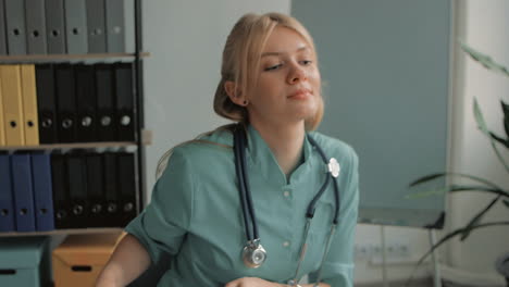 Junge-Hübsche-Blonde-Weibliche-Krankenschwester,-Die-Am-Schreibtisch-Arbeitet-Und-Stethoskop-Und-Klemmbrett-Aufhebt