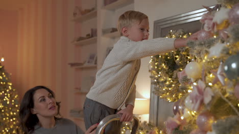 Junge-Schmückt-Weihnachtsbaum-Mit-Kugeln-Und-Ornamenten,-Seine-Mutter-Hilft-Ihm