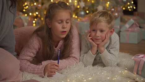 Kleines-Mädchen-Schreibt-Mit-Ihrem-Bruder-Einen-Brief-Auf-Dem-Boden-Neben-Den-Geschenken-Und-Dem-Weihnachtsbaum
