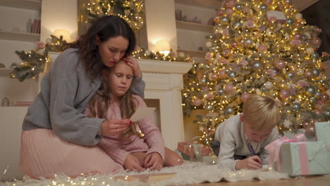 Kleiner-Junge-Und-Mädchen-Schreiben-Und-Lesen-Einen-Brief-Mit-Ihrer-Mutter-Auf-Dem-Boden-Neben-Den-Geschenken-Und-Dem-Weihnachtsbaum