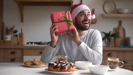 Excited-Man-Holds-Christmas-Present-While-Having-Breakfast-Wearing-Red-Santa-Hat