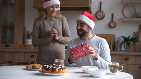 Young-Woman-Covers-Man-Eyes-To-Surprise-With-Christmas-Gift-At-Home