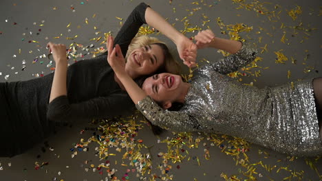 Two-Female-Friends-Lying-On-The-Floor-Full-Of-Confetti-After-A-Glamorous-New-Years-Eve-Party,-Laughing-And-Dancing-Hand-In-Hand