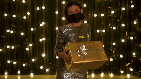 Young-Woman-With-Face-Mask-And-Evening-Dress-Holding-A-Golden-Gift-Box-And-Falling-Glitter-Confetti