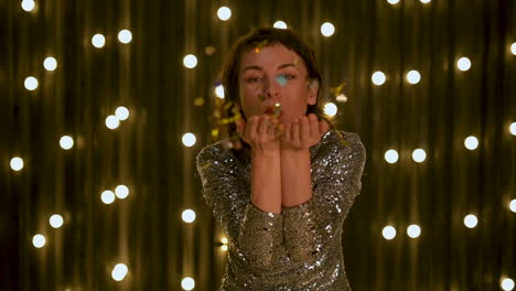 Young-Beautiful-Woman-Blowing-Gold-Glitter-Confetti-And-Looking-At-Camera