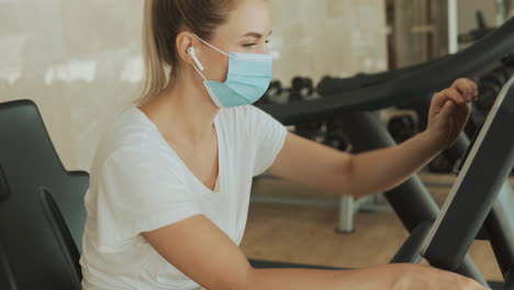 Young-Athlete-Female-With-Face-Mask-Uses-A-Smartphone-And-An-Exercise-Machine-In-The-Gym