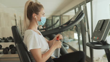 Young-Athlete-Female-With-Face-Mask-Uses-An-Exercise-Machine-And-Drinks-Water-In-The-Gym