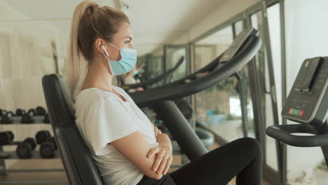 Young-Athlete-Female-With-Face-Mask-Uses-An-Exercise-Machine-And-Looks-To-Camera