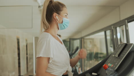 Young-Athlete-Female-With-Face-Mask-Uses-An-Exercise-Machine-And-Takes-A-Break-Breathing-Deep-In-The-Gym