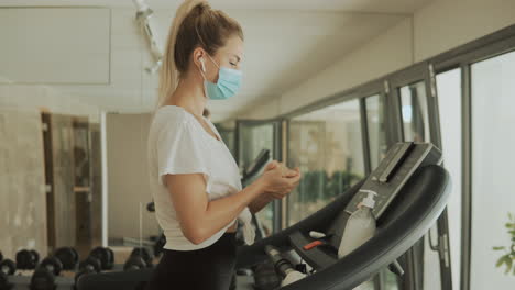 Young-Athlete-Female-With-Face-Mask-Uses-An-Exercise-Machine-And-Hand-Sanitizer-In-The-Gym