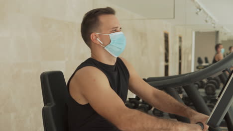 Young-Strong-Man-With-Face-Mask-Uses-A-Smartphone-And-An-Exercise-Machine-In-The-Gym
