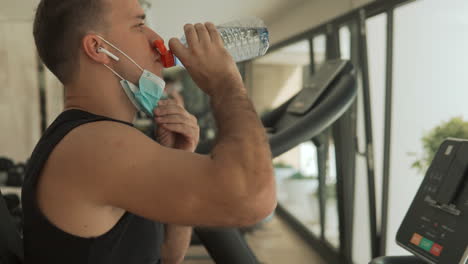Young-Strong-Man-With-Face-Mask-Uses-An-Exercise-Machine-And-Drinks-Water-In-The-Gym