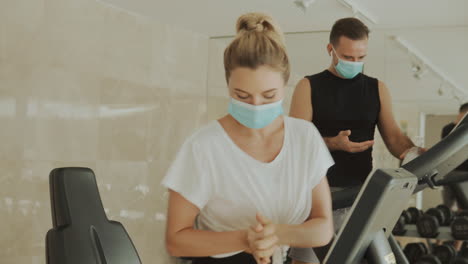 Young-Athlete-Female-And-Male-With-Face-Mask-Using-Hand-Sanitizer-And-Exercise-Machines-In-The-Gym