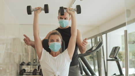 Young-Athlete-Woman-Lifts-Weights-And-Man-With-Face-Mask-Exercising-In-The-Gym