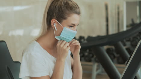Young-Sporty-Woman-Puts-On-Face-Mask-Ands-Uses-An-Exercise-Machine-In-The-Gym