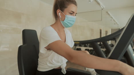 Athlete-Female-With-Face-Mask-Working-Out-With-An-Exercise-Machine-And-Drinking-Water-In-The-Gym
