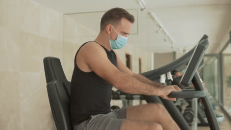 Strong-Male-With-Face-Mask-Uses-An-Exercise-Machine-And-Drinks-Water-In-The-Gym