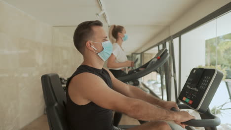Athlete-Male-And-Female-With-Face-Mask-Using-Exercise-Machines-In-The-Gym