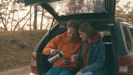 -Young-woman-and-man-sitting-in-the-open-trunk-of-the-car-having-a-hot-drink-with-a-thermos