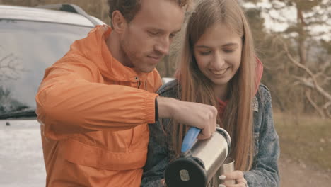 -Young-couple-sitting-on-the-car-having-a-hot-drink-with-a-thermos
