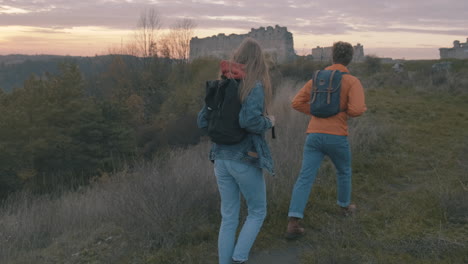 -Young-couple-of-female-and-male-hikers-at-the-top-of-the-mountain-find-ruins-of-an-ancient-castle-Beautiful-sunset
