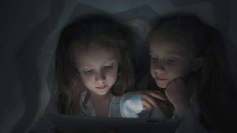 Two-Little-Girls-Using-A-Tablet-Covered-By-The-Bed-Sheet-At-Night