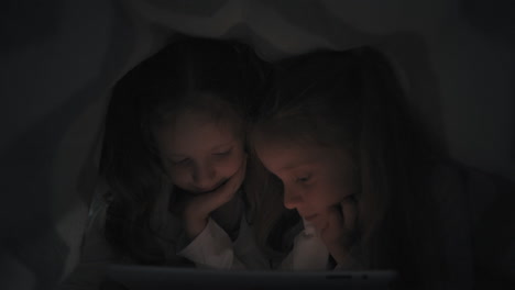 Two-Little-Sisters-Using-A-Tablet-Hiding-Under-The-Blanket-In-Bed-At-Night