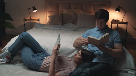 Young-Couple-Sitting-In-Bedroom,-Woman-And-Man-Reading-A-Tablet-And-A-Book-And-Relaxing,-Sharing-Moments