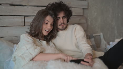 Young-Cute-Couple-Sitting-In-Bed-Watching-Something-Funny-On-The-Phone-And-Wearing-Winter-Clothes