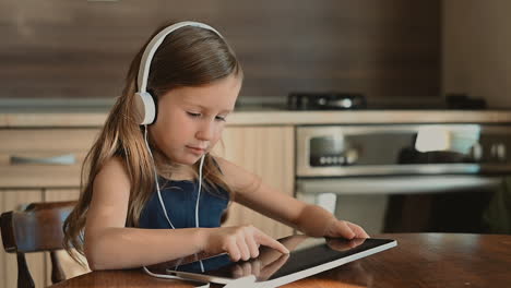 Little-Girl-With-Headphones-Uses-Tablet-At-Home