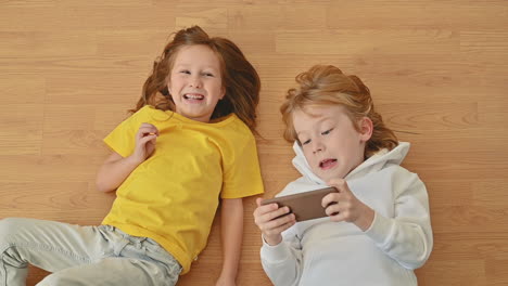 Kleines-Mädchen-Und-Junge-Mit-Einem-Smartphone,-Das-Auf-Dem-Boden-Liegt,-Eine-App-Benutzt-Und-Ein-Online-Videospiel-Spielt