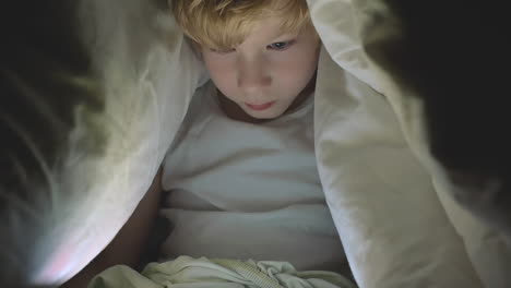 Little-Boy-Using-A-Tablet-Watching-A-Movie-Covered-By-The-Bed-Sheet-At-Night