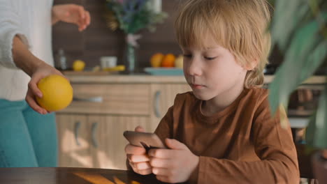 Little-Boy-Watching-Smartphone-At-Home,-Mother-Gives-Him-A-Fruit