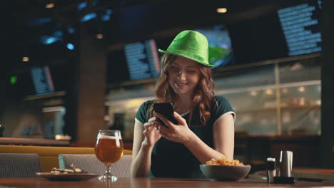 Hübsche-Junge-Frau,-Die-Ein-Smartphone-Benutzt,-Bier-Trinkt-Und-Einen-Irischgrünen-Hut-Trägt