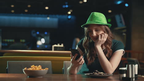 Hübsche-Junge-Frau,-Die-Auf-Jemanden-Wartet,-Besorgt,-Auf-Das-Telefon-Schaut-Und-Einen-Irisch-grünen-Hut-Trägt