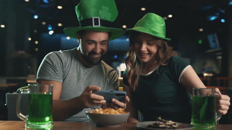 Joven-Y-Mujer-Con-Sombreros-Irlandeses-Con-Jarras-De-Cerveza-Verde-Tomando-Un-Selfie