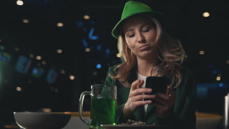 Porträt-Einer-Blonden-Frau,-Die-Ein-Smartphone-Benutzt,-Einen-Grünen-Bierkrug-Trinkt-Und-Einen-Irischen-Grünen-Hut-Trägt