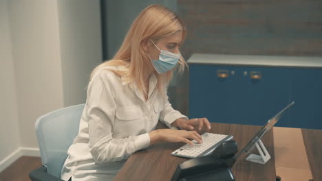 Young-Business-Female-With-Face-Mask-Typing-On-Tablet-In-The-Office