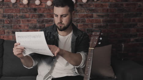 Young-Musician-Man-Looking-At-Sheet-Music-To-Play-Guitar-At-Home