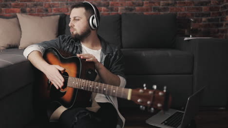 Young-Musician-Man-With-Guitar,-Listening-To-Music-With-Wireless-Headphones-And-Using-Laptop-At-Home