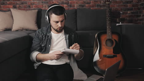 Young-Musician-Man-Listening-To-Music-With-Laptop-And-Wireless-Headphones-Taking-Notes-At-Home